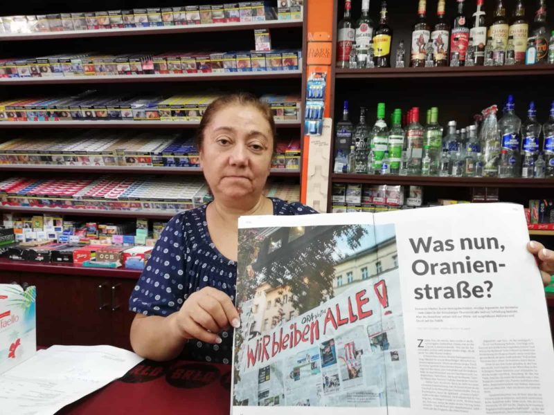 Frau in einem Späti hält Zeitung mit Aufschrift „Was nun, Oranienstraße?“ hoch